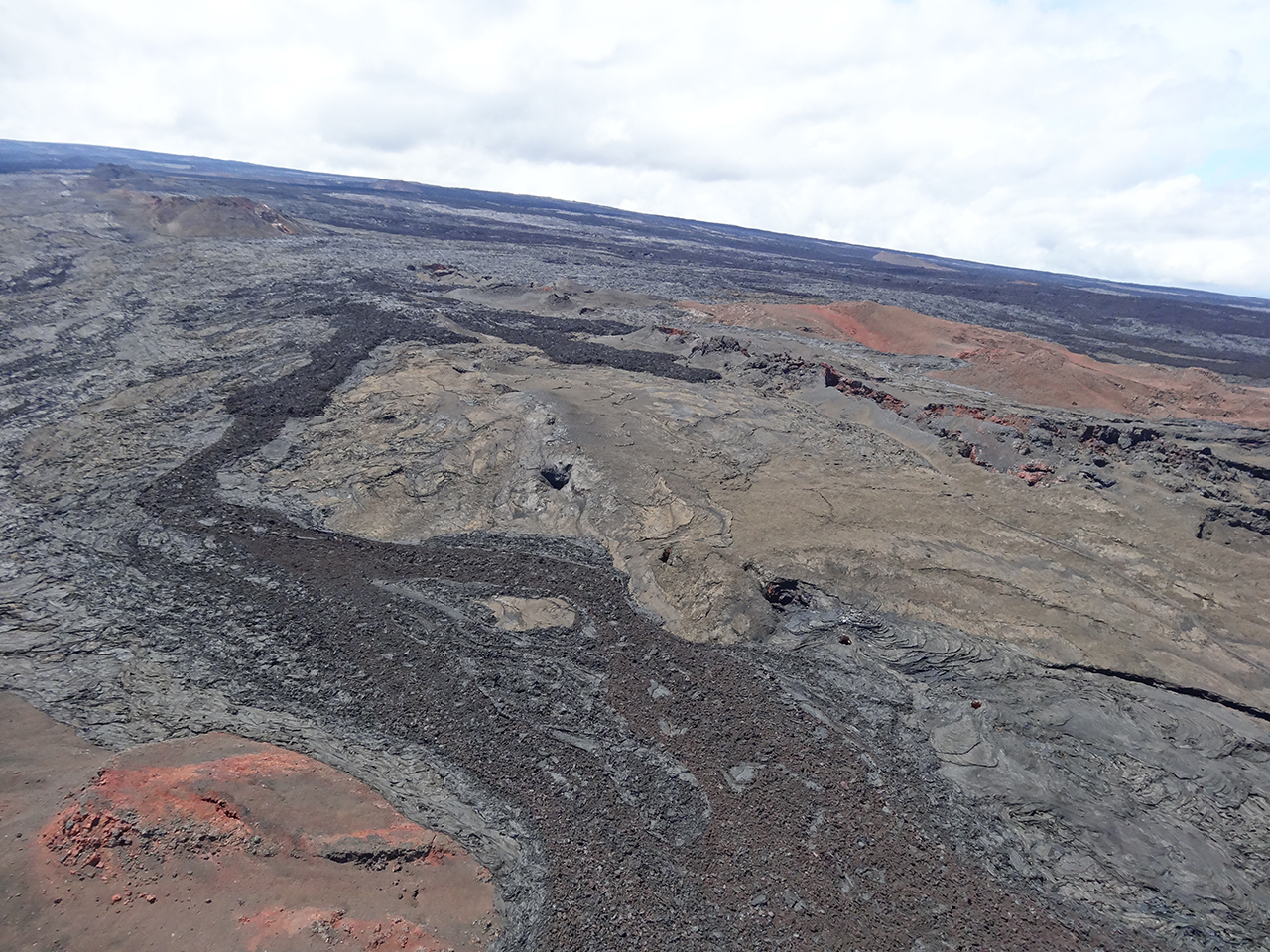 (USGS) Convergence of 3 historical eruptions: aa (dark flows) in the foreground and center are from the 1899 eruption.  Black phh lower right and tan center are flows from the 1880-81 eruption.  Beyond the 1880 is another fissure system in the center of the view (gray colored with red interiors) are from the 1855-56 eruption.  Prehistoric cone (tan-red) in theright central portion of the photo.  Dark flows beyond the preshistoric cone are mixed aa and phh from the 1899 eruption  View uprift.