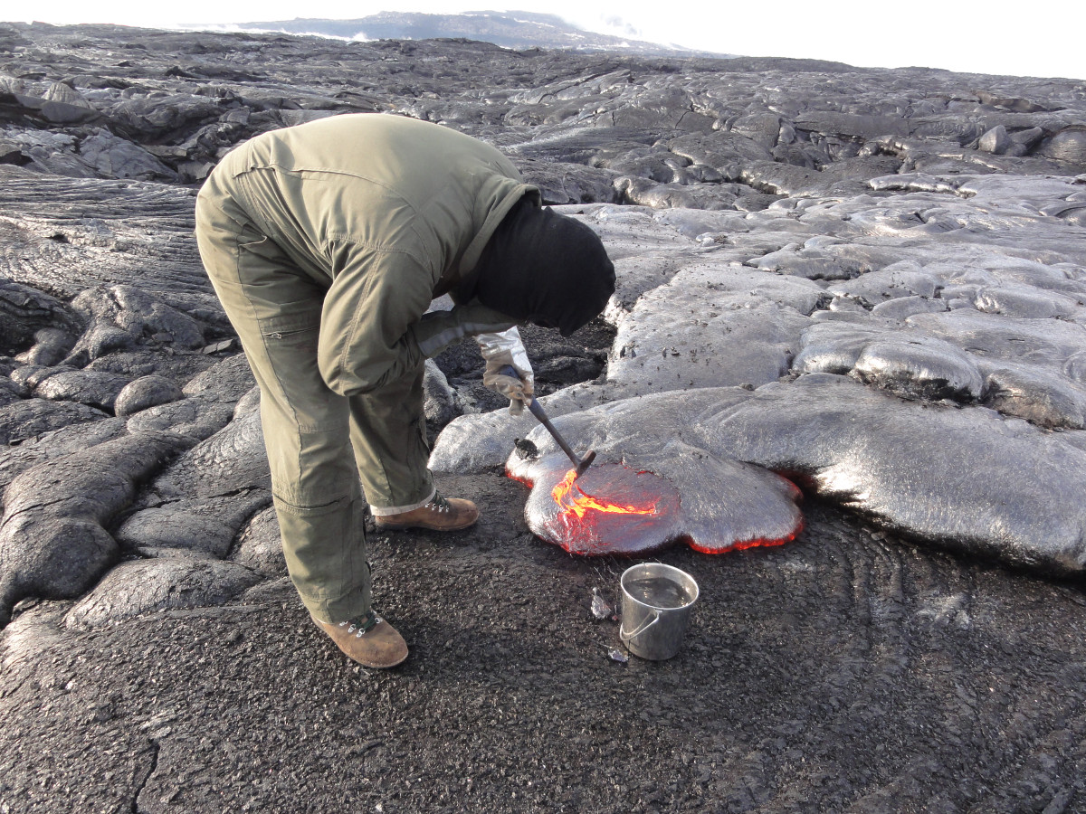 (USGS) An HVO geologist collects a molten lava sample for chemical analysis, scooping up a bit with the rock hammer to then drop in the water bucket to quench it. Puʻu ʻŌʻō is visible in the distance.