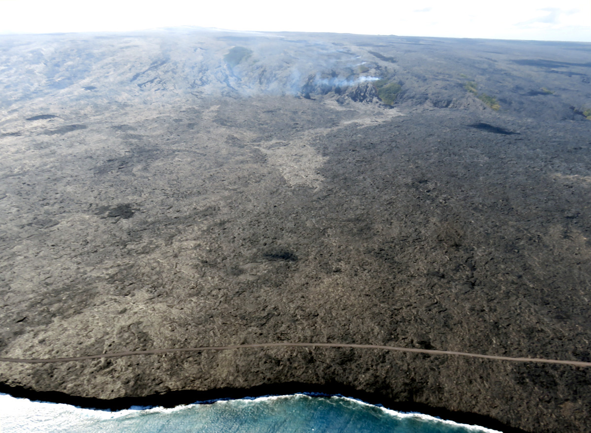 (USGS photo) The position of the lava flow front relative to the shoreline can be seen in this aerial photograph. The leading edge of the flow, which was 1.1 km (0.7 miles) from the ocean today, is the light-colored area near the center of the image. More vigorous breakouts were active upslope, near the base of the pali. Fume from the lava tubes and smoke from burning vegetation are visible on the pali in the upper part of the photo. Puʻu ʻŌʻō is visible on the upper left skyline.