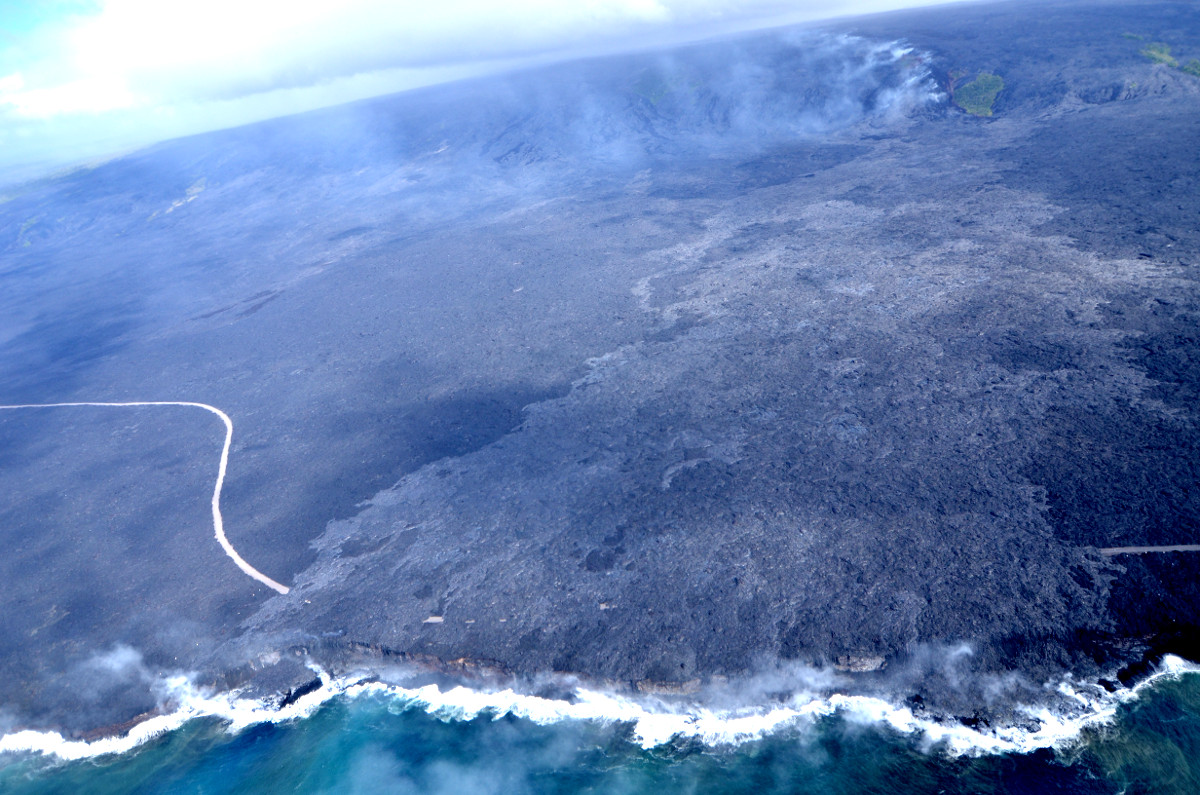 (USGS photo) Aerial view of the Kamokuna ocean entry and the Emergency Access Road cut by the 61g lava flow. 61g lavas are lighter in color than older lavas on the coastal plain. Upslope, a trail of fume marks the lava tube as it passes over the pali.