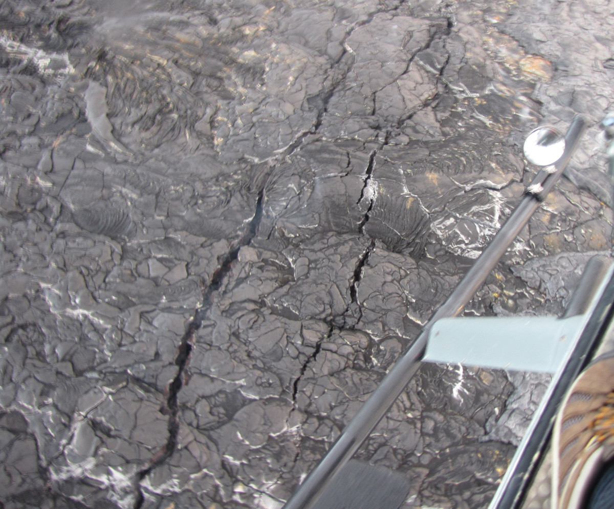 (Photo by Rick Hazlett, University of Hawaiʻi at Hilo) Looking down from the helicopter, cracks are visible on the surface of the east Kamokuna lava delta. These cracks are reminders that lava deltas are inherently unstable features that can collapse without warning. A lava delta collapse can send tons of hot rock into the sea, generating steam-driven explosions that can hurl fragments of molten lava and solid rock 100s of meters (yards) in all directions—inland and seaward. 