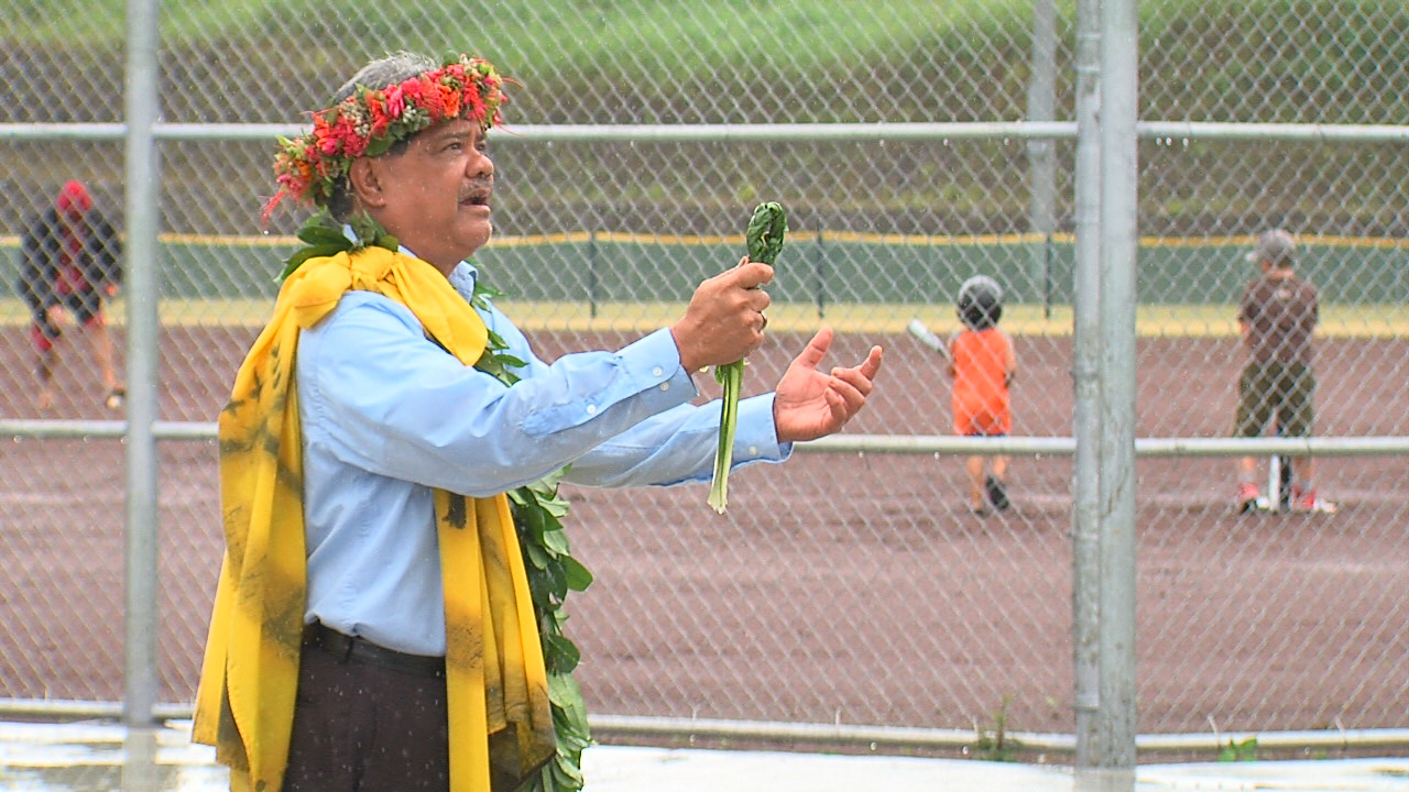 Standing in the rain, Kumu Crabbe blesses the new facilities in Pahoa, while a family plays on the baseball field behind him.